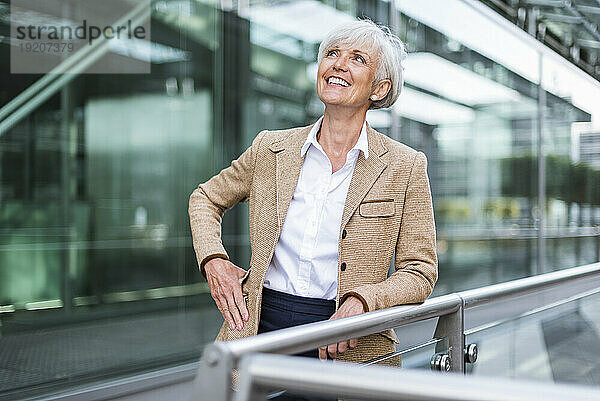 Lächelnde ältere Geschäftsfrau lehnt am Geländer in der Stadt und schaut nach oben