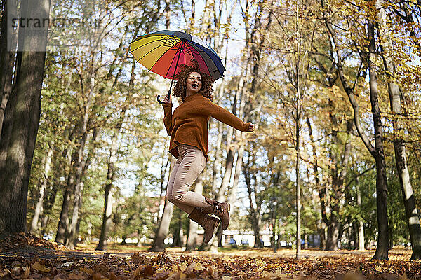 Lächelnde rothaarige Frau springt mit buntem Regenschirm im Herbstpark