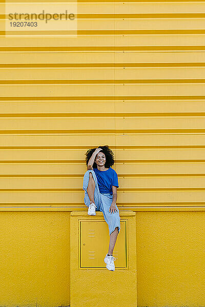 Hübsche Frau sitzt lächelnd auf einem Bahnsteig vor einer gelben Wand
