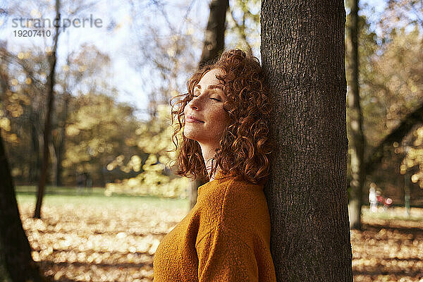 Rothaarige Frau mit geschlossenen Augen lehnt an Baum im Herbstpark