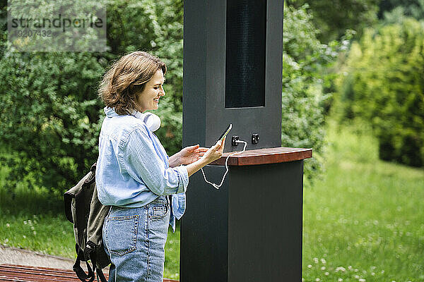 Lächelnde Frau lädt Smartphone durch Solarenergie im Park auf