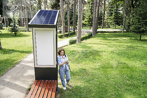 Frau surft mit Smartphone im Internet in der Nähe von Solarladestation im Park