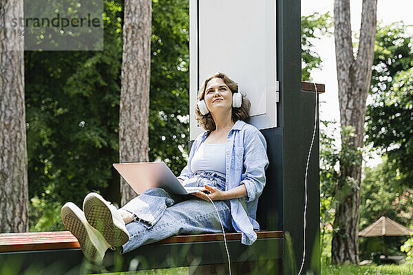 Lächelnde Frau träumt davon  mit Laptop an der Solarladestation im Park zu sitzen