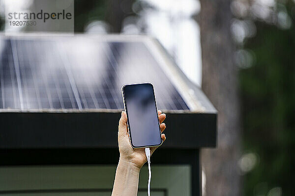Hand einer Frau  die ein Smartphone mit Solarpanel im Hintergrund hält