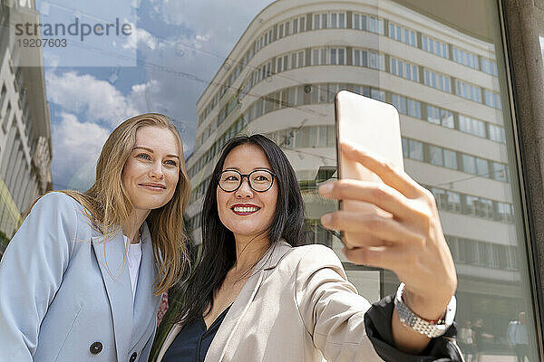 Lächelnde Geschäftsfrau macht Selfie mit Kollege in der Nähe einer Glaswand