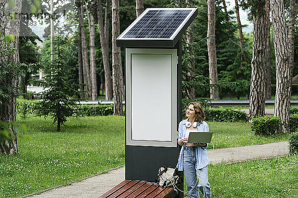 Frau mit Laptop steht in der Nähe einer Solarladestation im Park