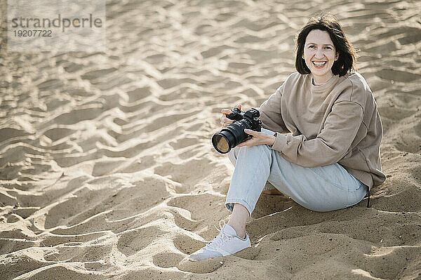 Glückliche Frau sitzt mit Kamera im Sand am Strand