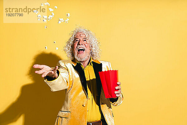 Fröhlicher älterer Mann fängt Popcorn vor gelbem Hintergrund