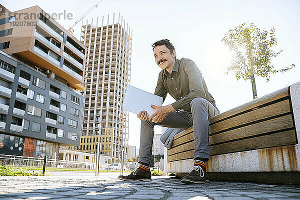 Lächelnder  nachdenklicher Geschäftsmann mit Laptop  der auf einer Bank im Büropark sitzt