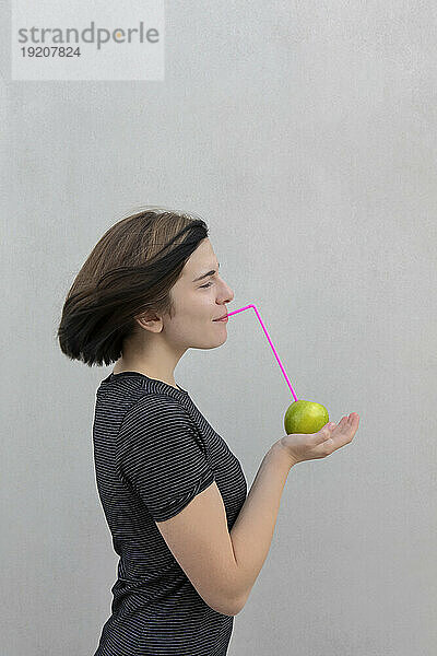 Teenager-Mädchen hält Granny-Smith-Apfel mit Stroh vor grauem Hintergrund