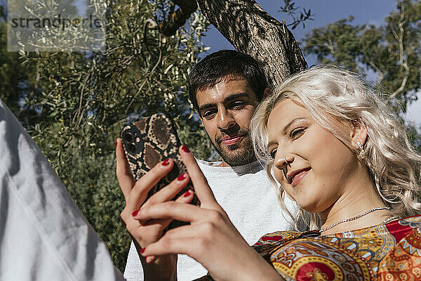 Lächelnde Freundin zeigt und benutzt Smartphone mit Freund vor Baum
