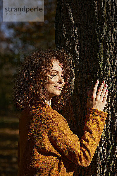 Lächelnde rothaarige Frau umarmt Baum im Herbstpark