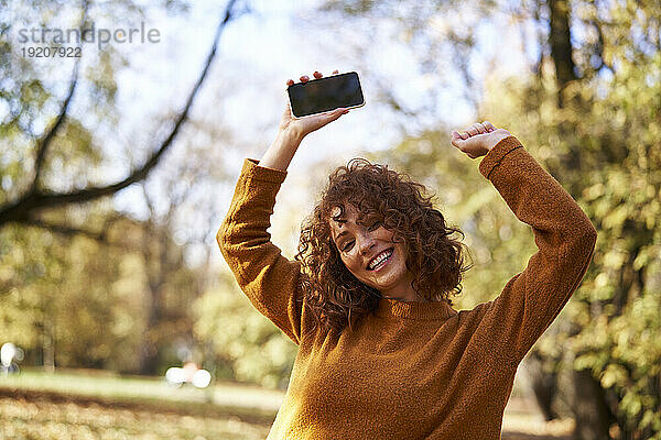 Fröhliche rothaarige Frau tanzt mit Smartphone im Herbstpark