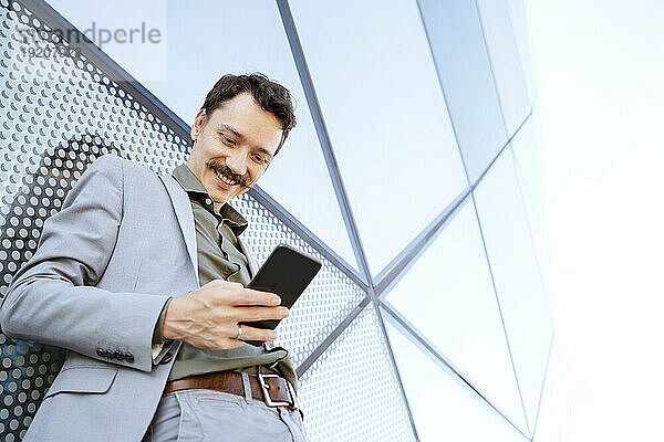 Lächelnder Geschäftsmann mit Smartphone vor modernem Glasgebäude