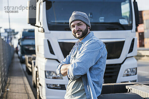 Glücklicher LKW-Fahrer mit verschränkten Armen  der an einem sonnigen Tag vor dem LKW steht