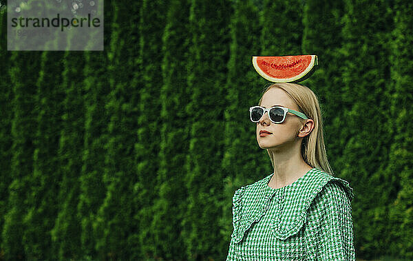 Frau mit Sonnenbrille steht mit Wassermelone auf dem Kopf im Garten