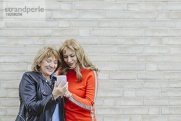 Lächelnde Mutter und Tochter schauen vor einer Ziegelwand auf ihr Smartphone