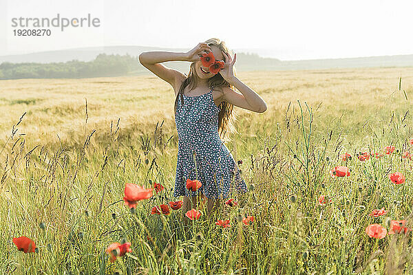 Mädchen hält an einem sonnigen Tag im Feld rote Mohnblumen über die Augen