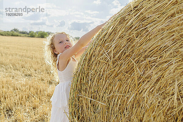 Blondes Mädchen berührt an einem sonnigen Tag einen Strohballen auf dem Feld