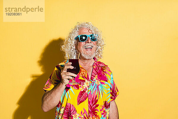 Fröhlicher älterer Mann mit einem Glas Saft vor gelbem Hintergrund