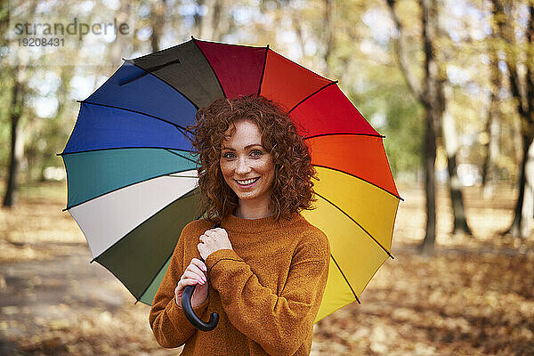Smiling redhead woman holding colorful umbrella at autumn park