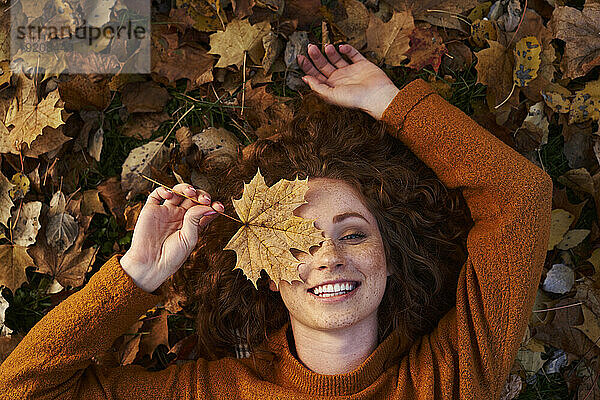 Smiling redhead woman holding maple leaf and lying on ground at park