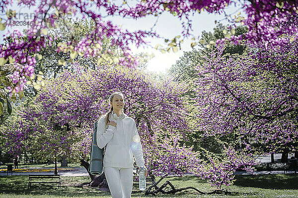 Teenage girl carrying exercise mat and water bottle walking at park in bloom