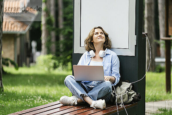Nachdenkliche Frau mit Laptop sitzt in der Nähe einer Solarladestation im Park