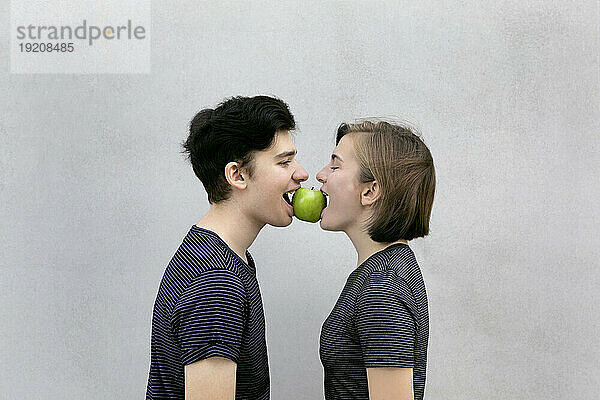 Teenager-Paar trägt gemeinsam Apfel im Mund vor grauem Hintergrund