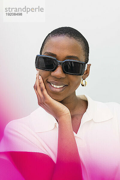 Happy woman wearing sunglasses standing with hand on chin in front of white wall