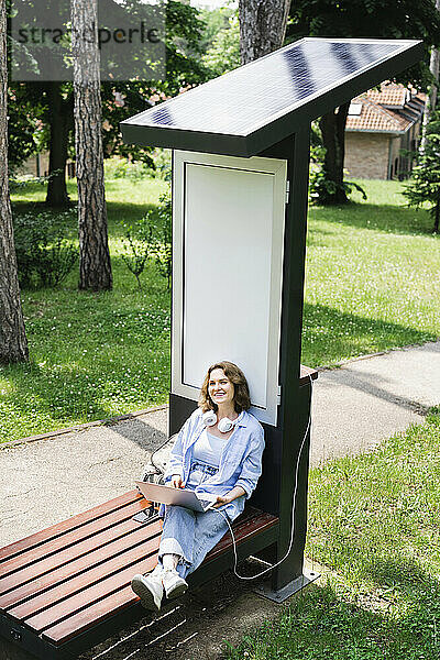 Lächelnde Frau mit Laptop sitzt auf einer Bank in der Nähe der Solarladestation im Park