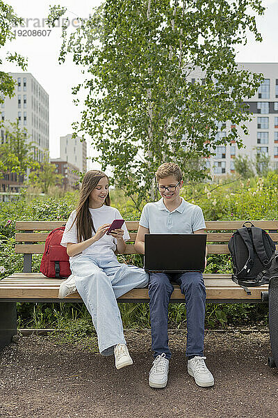 Friends using smart phone and laptop sitting on bench at park