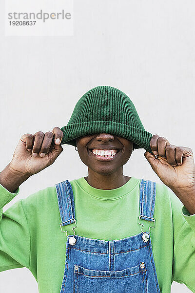 Happy woman covering face with green knit hat in front of white wall
