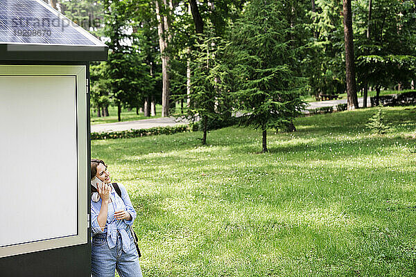 Frau telefoniert mit Handy und lehnt an Solarladestation im Park