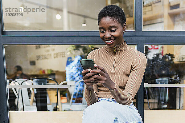 Glückliche junge Frau benutzt Mobiltelefon vor Glasfenster