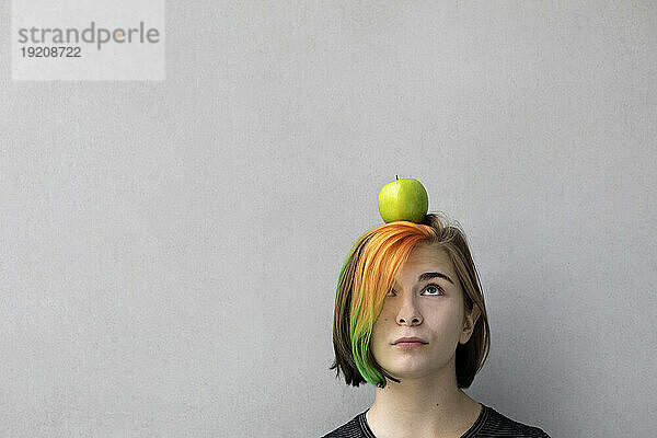 Teenager-Mädchen mit gefärbten Haaren balanciert Apfel auf dem Kopf vor grauem Hintergrund