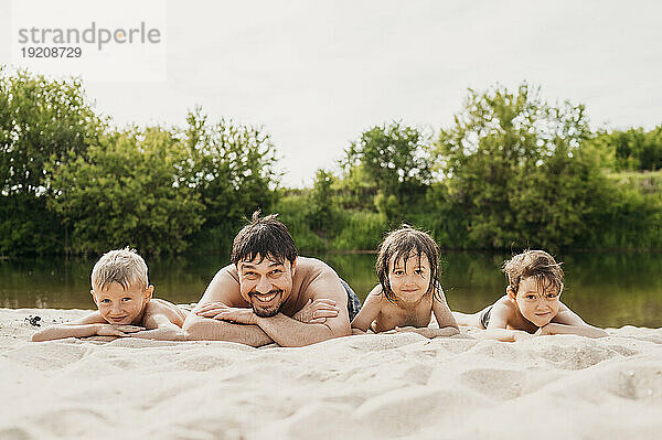 Vater liegt mit Kindern am Flussufer im Sand