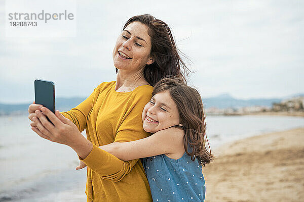 Lächelnde Frau macht mit ihrer Tochter am Strand ein Selfie per Smartphone