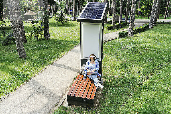 Frau sitzt mit Laptop auf Bank in der Nähe von Solarladestation im Park