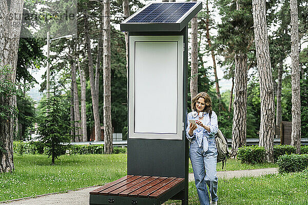 Frau benutzt Mobiltelefon und steht in der Nähe einer Solarladestation im Park