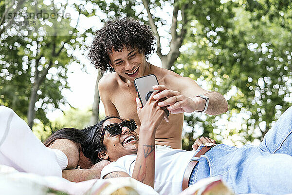 Lächelnde Freundin liegt auf dem Schoß ihres Freundes und schaut im Park auf ihr Smartphone