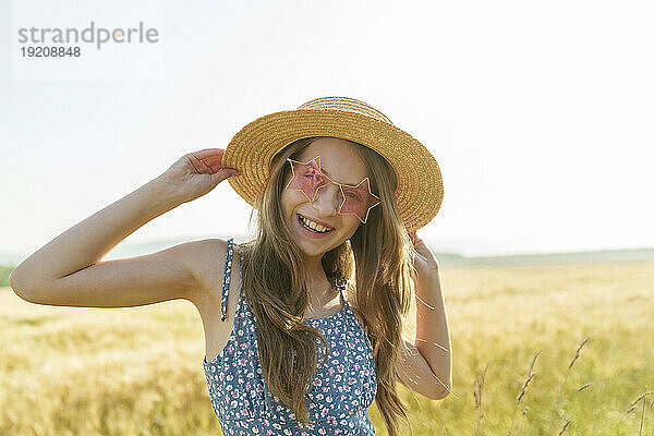 Lächelndes Mädchen mit sternförmiger Sonnenbrille und Hut auf dem Feld