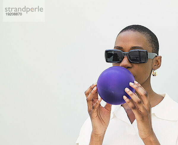 Young woman wearing sunglasses blowing balloon against white background