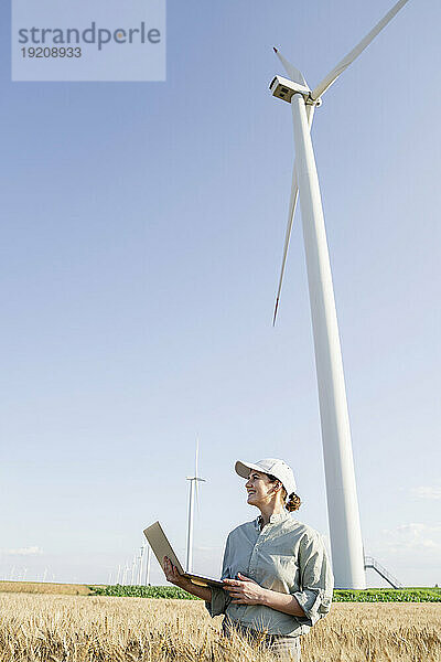 Glücklicher Bauer mit Laptop  der an einem sonnigen Tag vor Windkraftanlagen steht