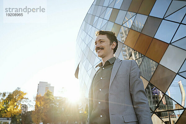 Lächelnder Geschäftsmann im Anzug  der an einem sonnigen Tag neben einem modernen Bürogebäude steht