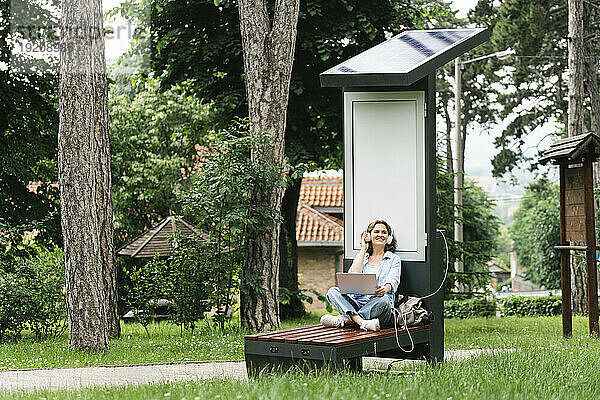 Frau sitzt auf einer Bank und hört Musik in der Nähe einer Solarladestation im Park