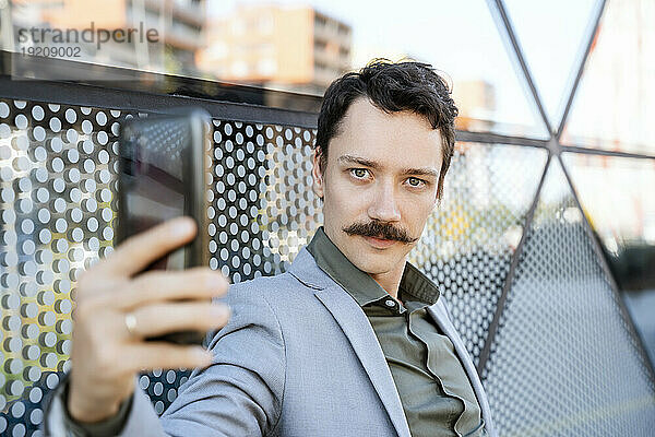 Geschäftsmann mit Schnurrbart macht Selfie mit Smartphone vor modernem Bürogebäude