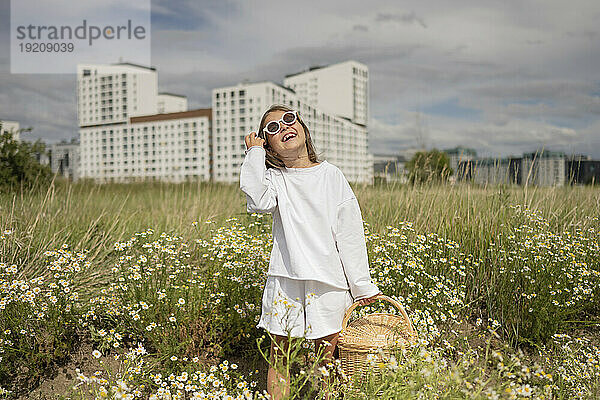 Lächelndes Mädchen mit Sonnenbrille hält Weidenkorb auf der Wiese