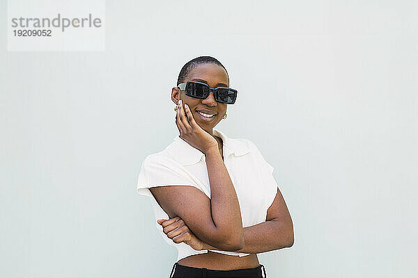 Smiling young woman wearing sunglasses and standing with hand on chin against white background