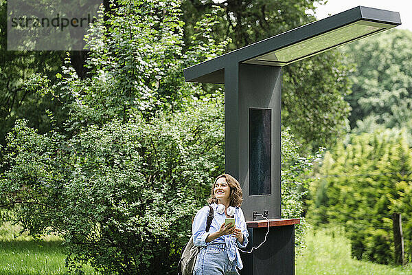 Lächelnde Frau steht in der Nähe einer Solarladestation im Park